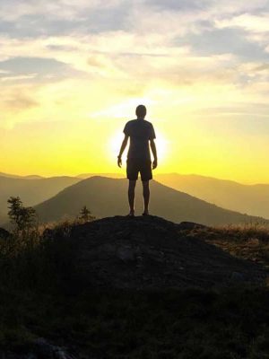 Silhouette of a person standing on a rock, overlooking a mountainous landscape at sunset. The sky is filled with glowing hues of orange and yellow, and soft clouds are scattered above.