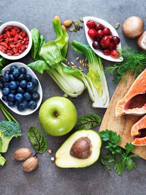A vibrant assortment of nutrition-packed fresh foods on a gray surface, featuring salmon fillets, avocado, berries, bok choy, apple, pomegranate, mushrooms, green beans, broccoli, and assorted leafy greens and herbs—all essential for a healthy brain.