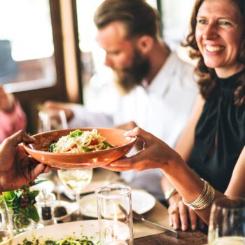 People enjoying a meal at a table, with a person passing a dish of pasta to another. A woman is smiling, and there are glasses of white wine and a candle on the table. The setting radiates warmth and social congruence, perfect for some weekend activation.