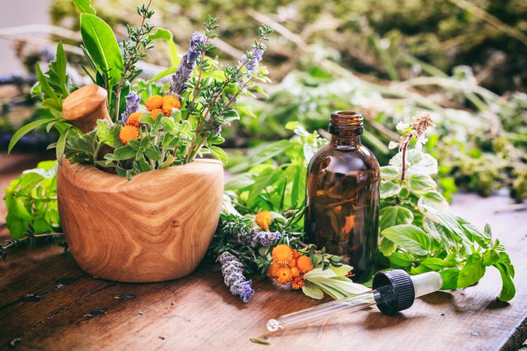 A wooden mortar filled with fresh herbs, small flowers, and flower essences sits on the table. Nearby are an amber dropper bottle, a pipette, and scattered botanicals—creating a natural apothecary scene reminiscent of Linda L. Gunter's approach to Neurosculpting.