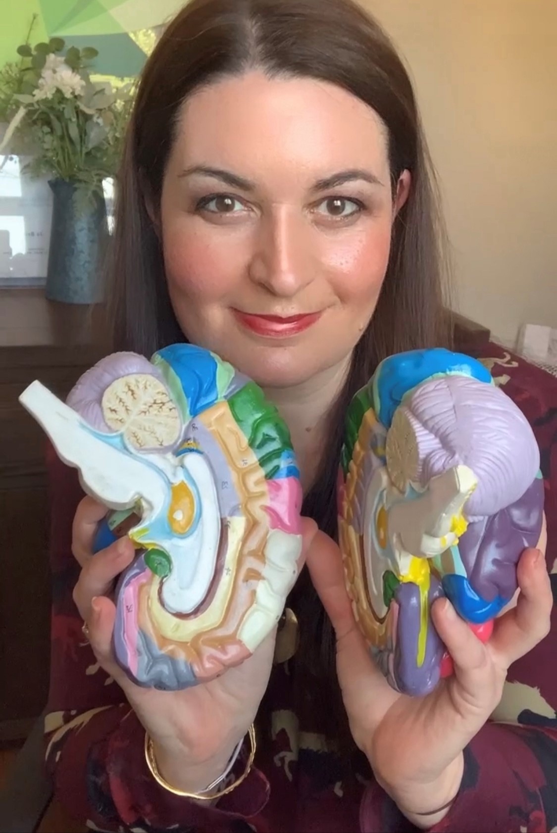 A woman with long brown hair holds two colorful cross-section models of the human brain, smiling at the camera. A vase with flowers and a green painting are visible in the background.