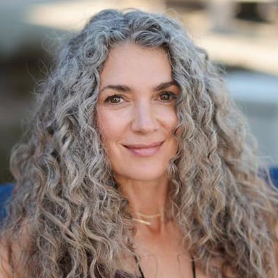 A woman with long, curly gray hair and a gentle smile looks directly at the camera. She stands outdoors with a blurred background, embodying the essence of a beginner on her Neurosculpting learning path.