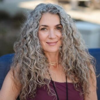 A person with long, curly gray hair smiles at the camera, exuding a sense of congruence. They wear a purple top and a necklace, sitting outdoors on a blue cushion. The background is beautifully out of focus, capturing the essence of this serene moment.
