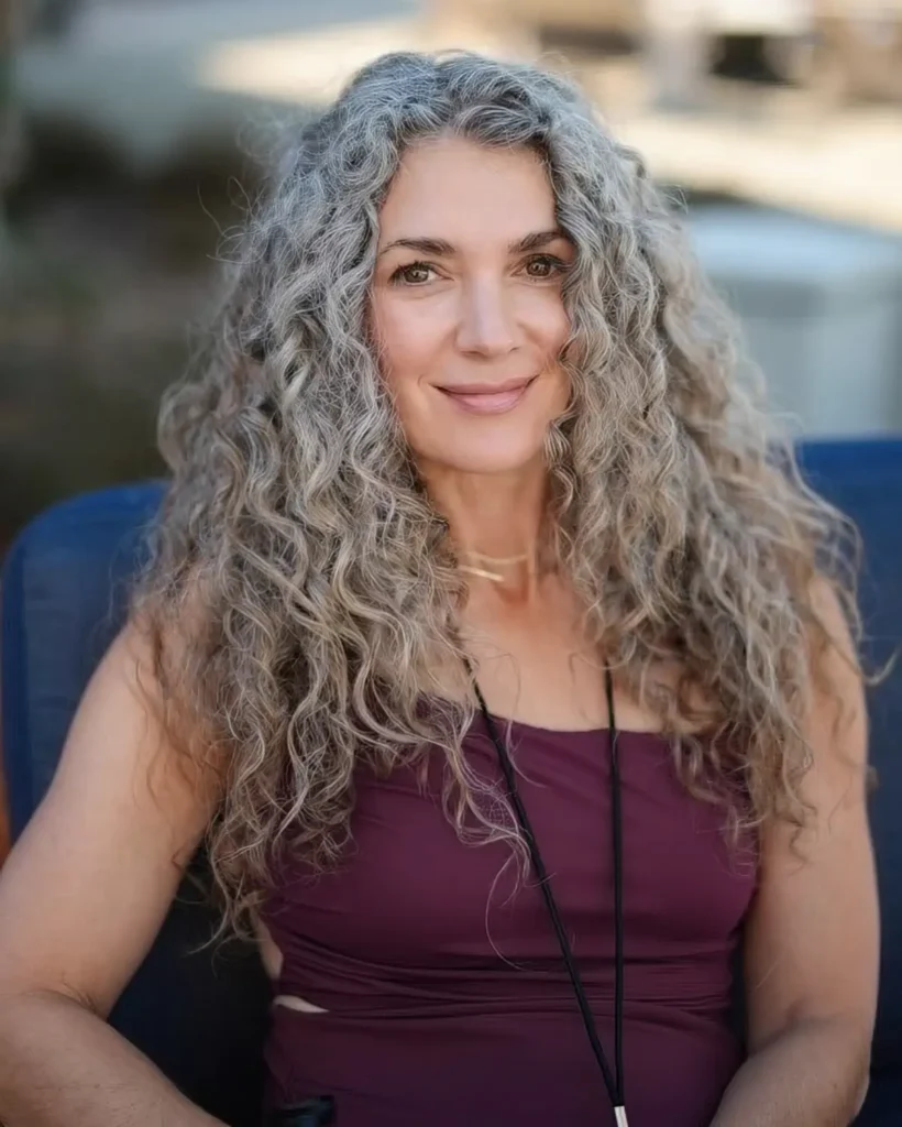 A woman with long, curly gray hair and a warm smile sits on a blue chair. She is wearing a sleeveless maroon top and a black necklace, with an outdoor background softly blurred behind her.