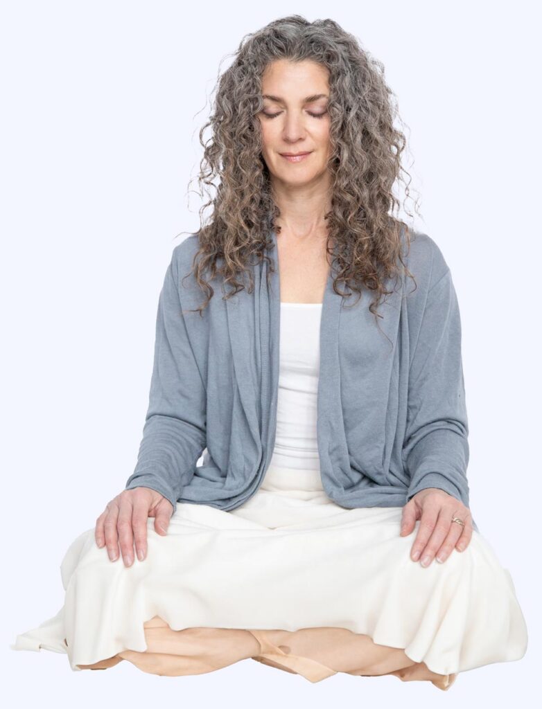A person with long curly hair sits cross-legged, deep in meditation. Clad in a light blue cardigan over a white outfit, their eyes are closed, exuding a sense of calm. The serene background reflects the soft hues of light blue.