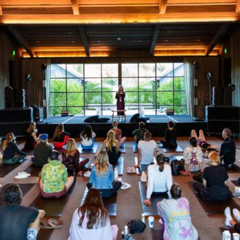 A group of people sits on yoga mats in a spacious, wooden room with large windows. They are facing a speaker on an elevated stage, embarking on a weekend activation journey. The room is well-lit with natural light from outside. The atmosphere is calm and focused, fostering deep transformation.