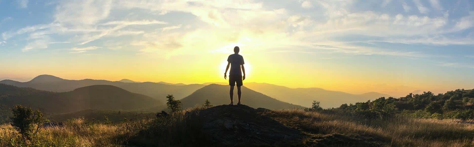 A person stands on a hilltop, silhouetted against a vibrant sunrise, embracing a mindset of wonder. The sky is painted with masterclass shades of orange and yellow, casting light over distant mountains and grassy foreground.