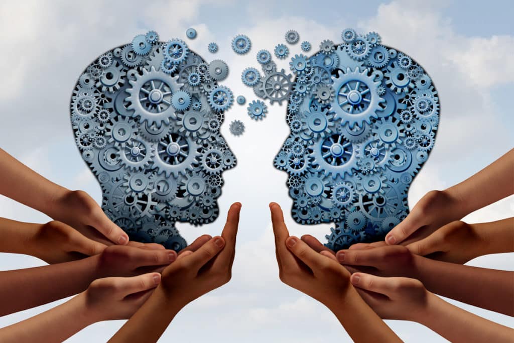 Illustration of two interconnected human profiles made of blue gears facing each other, symbolizing a beginner's learning path. Multiple hands support the profiles from below, emphasizing collaboration and the exchange of ideas against a cloudy sky background.