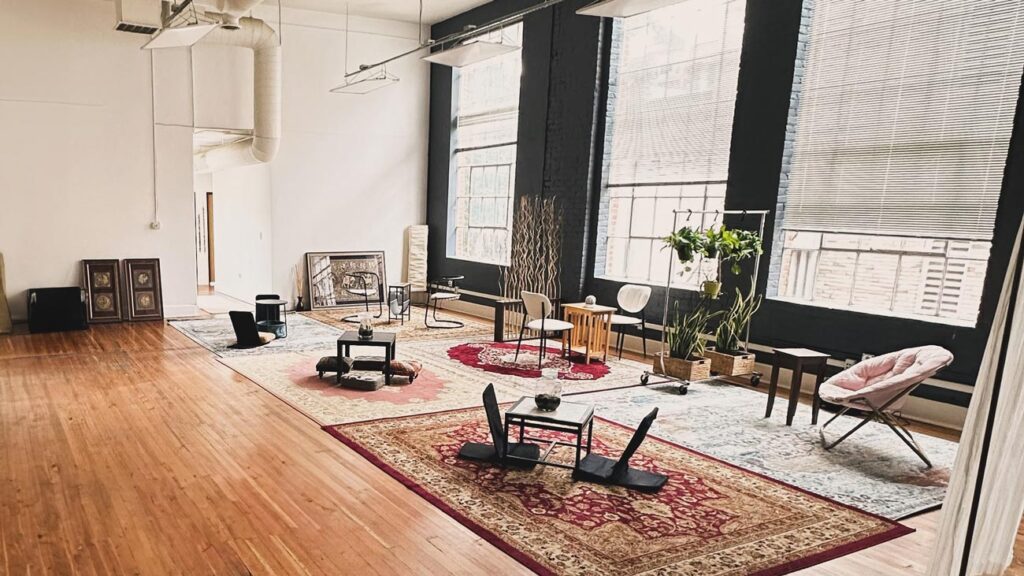 A spacious room with wooden flooring and high ceilings, featuring large windows with blinds. Several rugs are spread across the floor, each with low tables and floor seating. Potted plants and other small furnishings decorate the space.