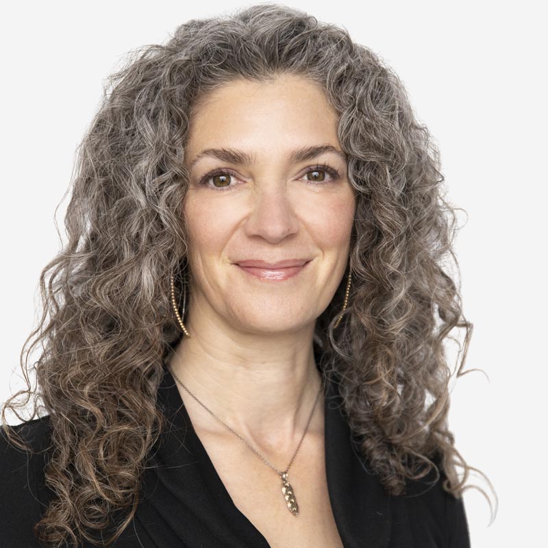 Woman with long curly gray hair smiles at the camera. She wears a black top and a necklace, posing against a plain white background. Her serene expression seems to unlock hope, inviting you to reshape your future with positivity and grace.