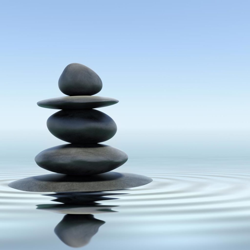A stack of smooth stones balances on a tranquil water surface, creating gentle ripples and inviting a sense of stress release. The sky is clear and blue, reflecting a calming atmosphere.