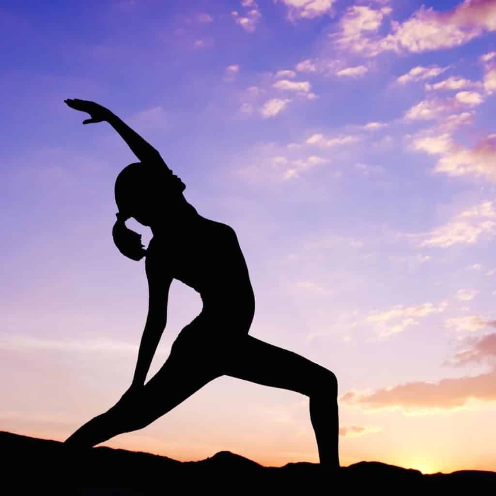 A silhouette of a person engaging in yoga graces the vibrant sunset sky, with colorful clouds casting their hues over distant hills, embodying the art of neurosculpting in perfect harmony.