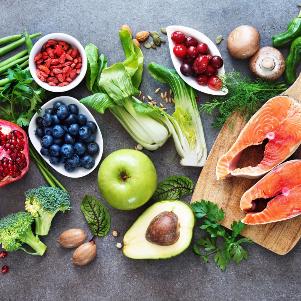 A vibrant assortment of nutrition-packed fresh foods on a gray surface, featuring salmon fillets, avocado, berries, bok choy, apple, pomegranate, mushrooms, green beans, broccoli, and assorted leafy greens and herbs—all essential for a healthy brain.