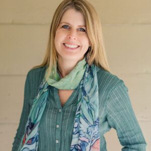 A woman with long blonde hair, wearing a green button-up blouse and a colorful scarf, stands smiling in front of a light beige wall.