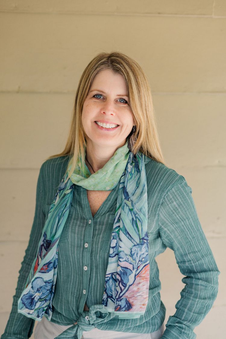 A woman with long blonde hair, wearing a green button-up blouse and a colorful scarf, stands smiling in front of a light beige wall.