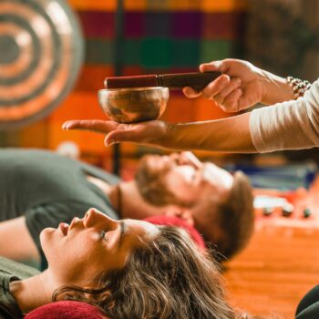Two people lie down with eyes closed, immersed in a transformative sound therapy session. A hand gently holds a singing bowl above them in the warm-toned, colorfully decorated room. A gong, symbolizing harmony and congruence, stands prominently in the background.