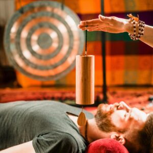 A person lies on a mat with eyes closed, immersed in deep transformation, while another person's hand holds a wooden chime above them. A large gong and colorful patterned fabric are in the background, suggesting a weekend activation or meditation session.