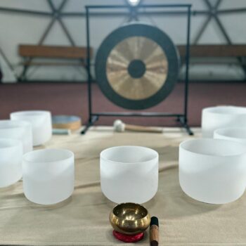 A collection of white crystal singing bowls is arranged on a table, with a brass singing bowl and mallet in the foreground, inviting a deep transformation. A large gong stands in the background, supported by a stand in this spacious indoor setting, perfect for weekend activation.