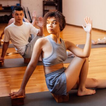 In a peaceful yoga studio, two people embody deep transformation through seated twists. The foreground person, in a gray tank top and shorts, sits on a block with serene focus. Behind them, another in white and gray absorbs the sunlight streaming in—a perfect weekend activation of mind and body.