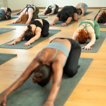 People are practicing yoga in a studio, all positioned on mats in child's pose. The wooden floor and soft lighting create a warm, calming atmosphere, fostering deep transformation for everyone involved.