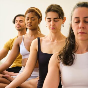 Four people sit cross-legged in a yoga class, eyes closed in meditation. Dressed in casual workout attire, they embody congruence as they appear calm and focused. The serene, well-lit setting enhances this deep transformation during their practice.